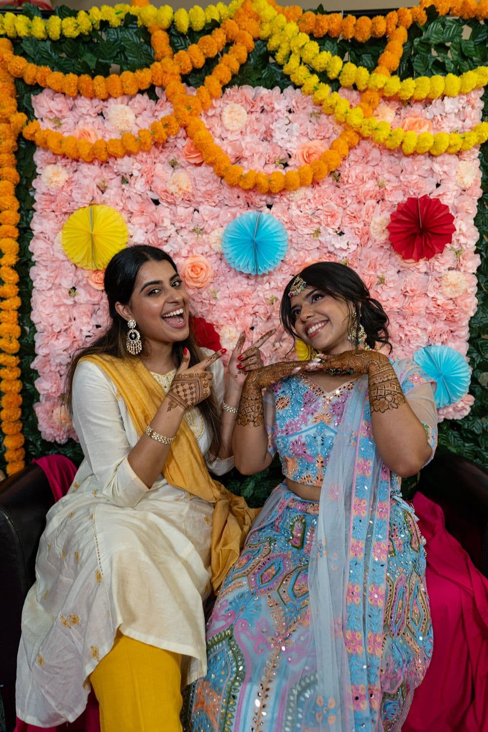 Two women smiling at a colourful mehndi night portrait setup.