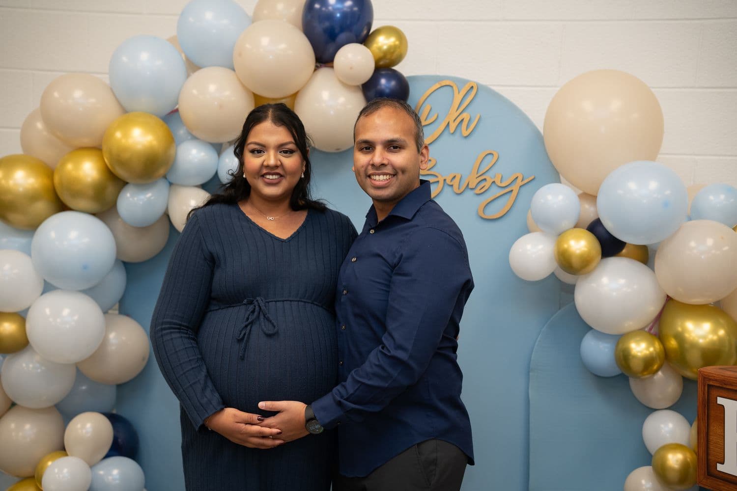 A couple posing in front of a blue and gold baby shower balloon display.