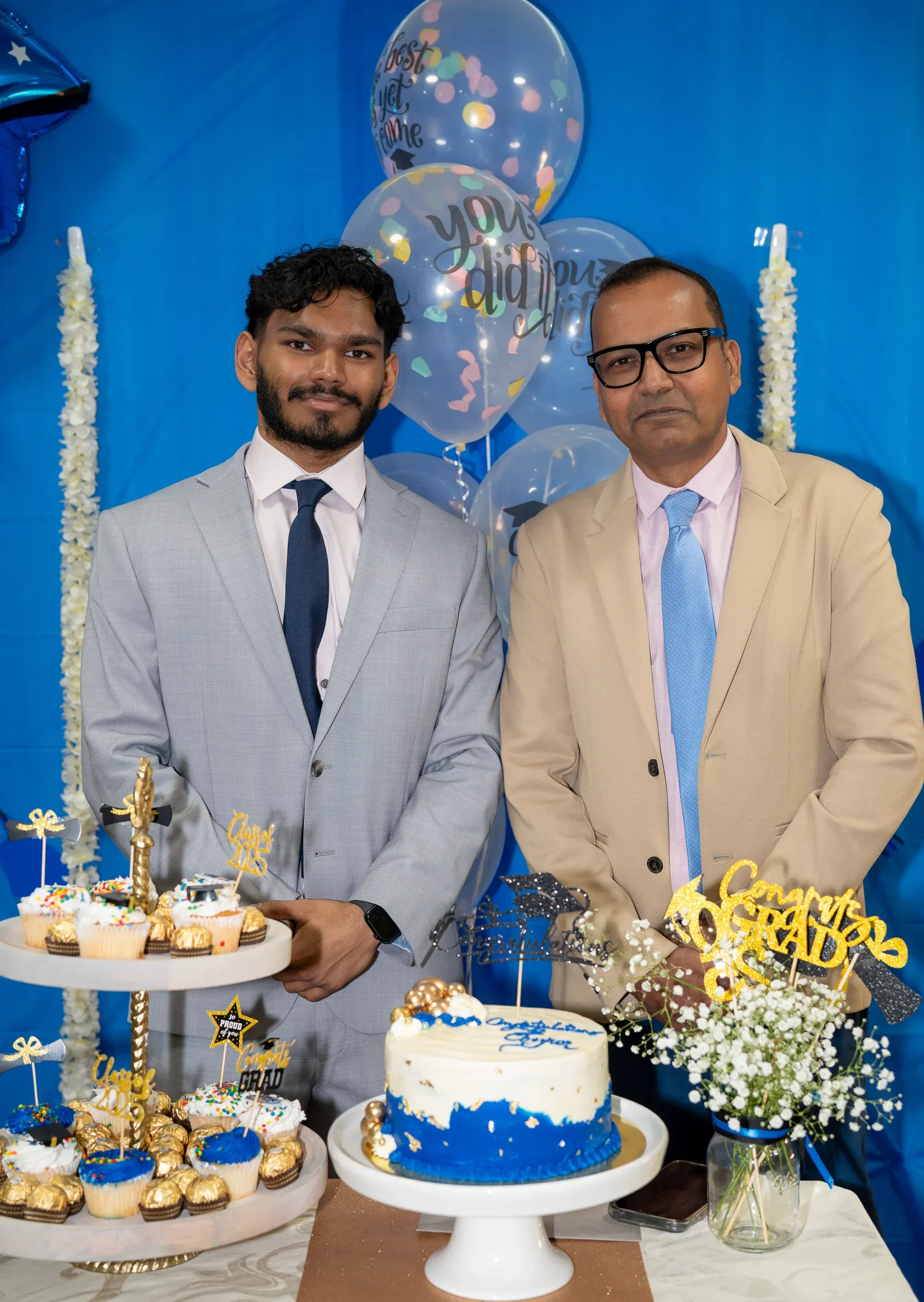 A graduate and his father cutting a celebration cake under blue and gold balloons.