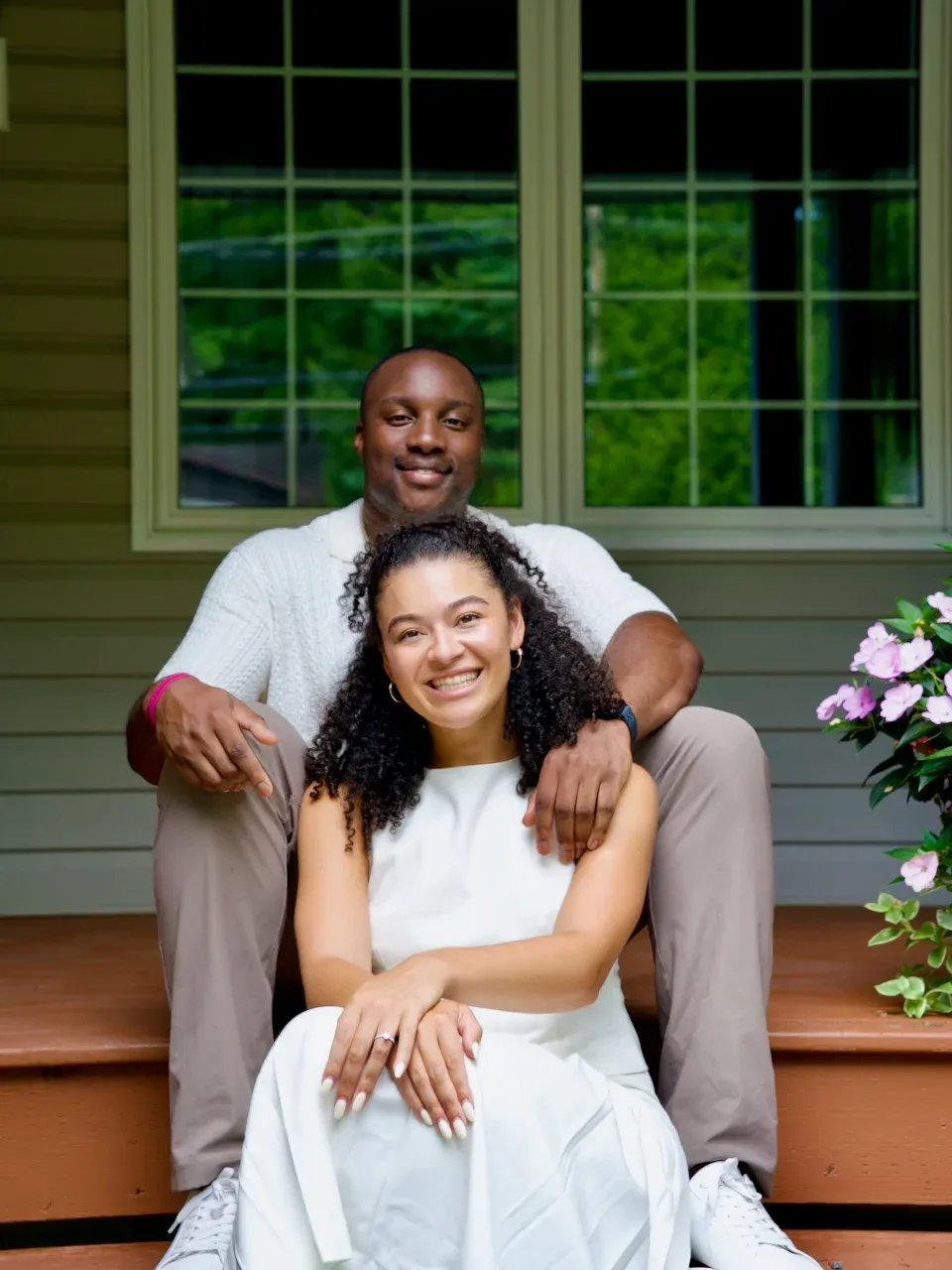 An engaged couple in white sitting close together on porch steps, the ring visible, summer greenery behind them.