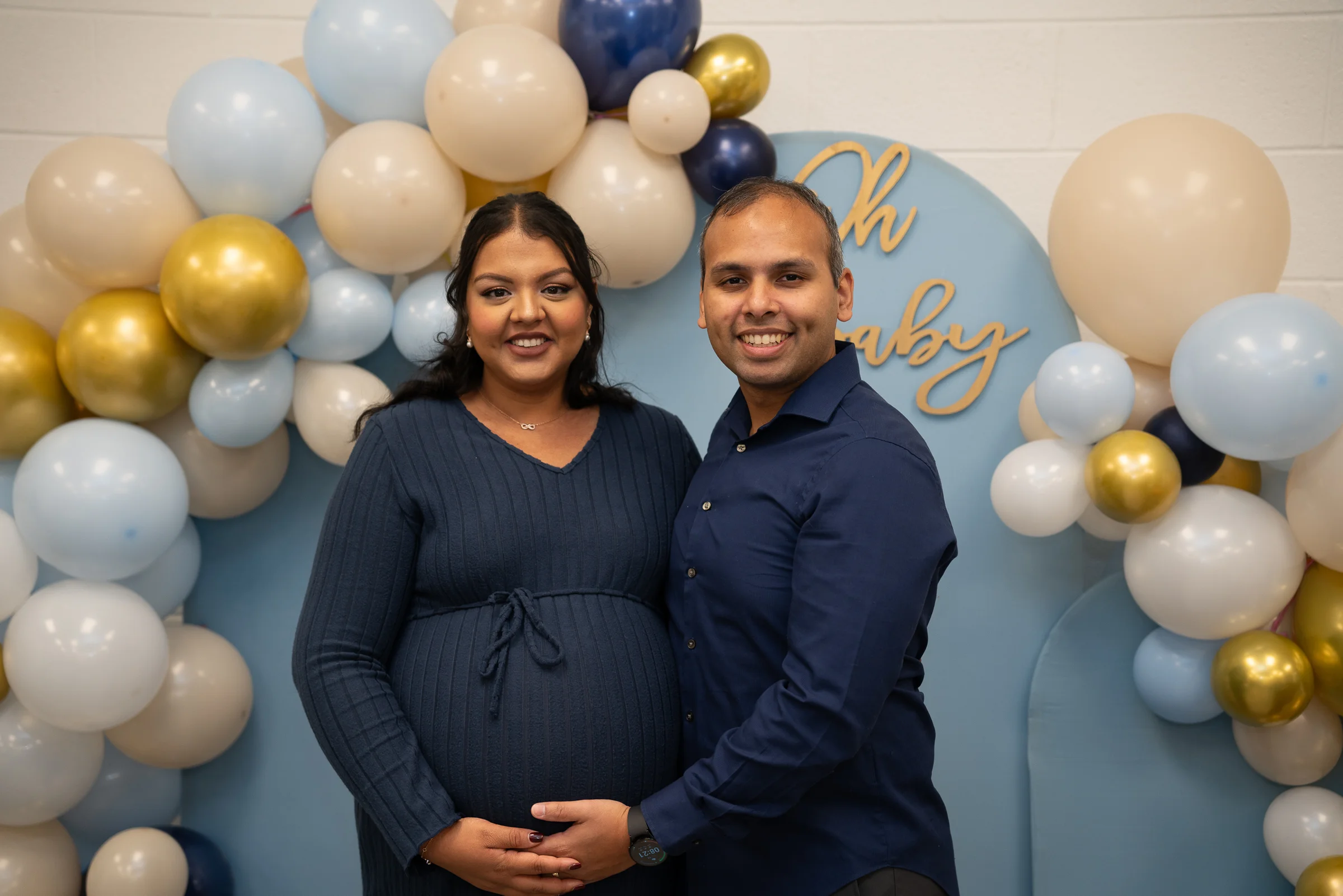 An expecting couple in navy standing together in front of a blue, white and gold balloon arch, hands resting on the bump.