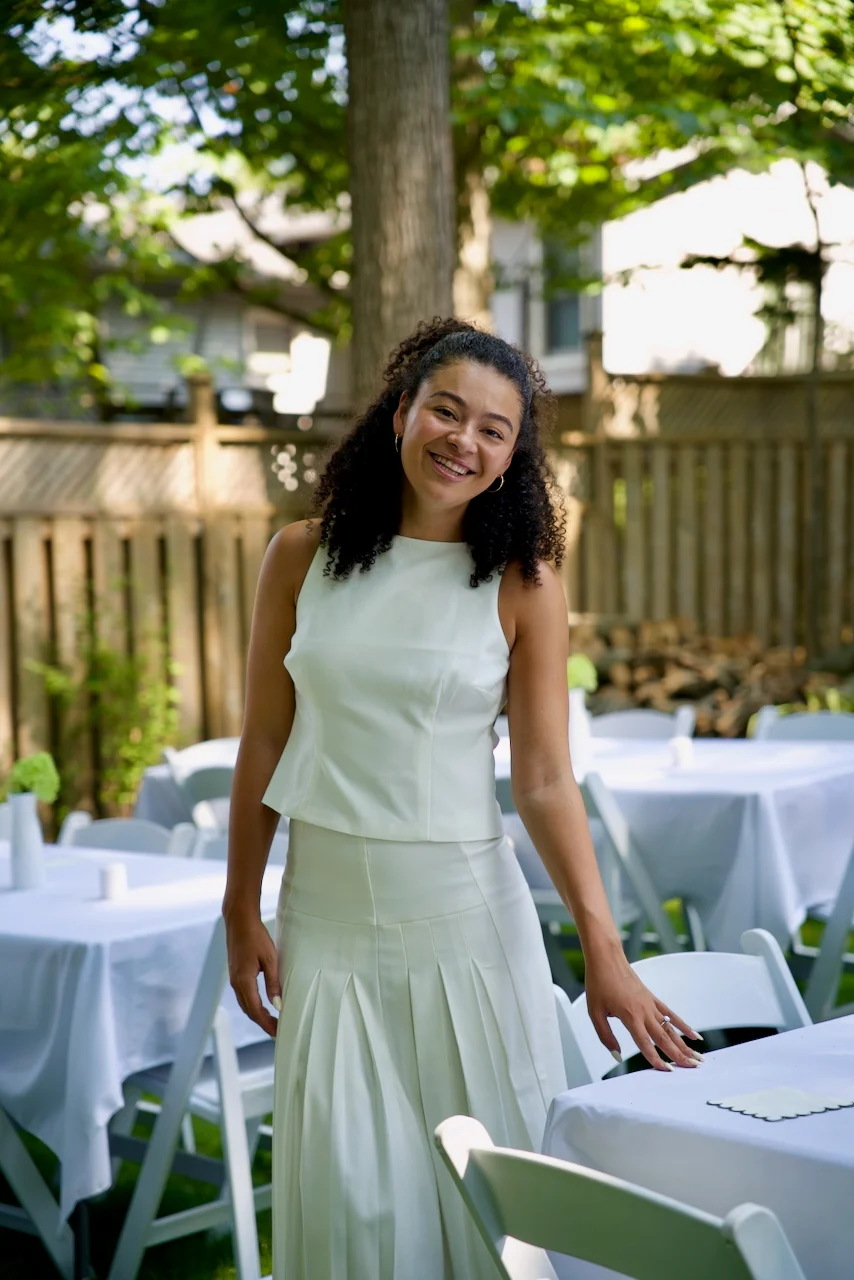 A woman in a white dress smiling in a sunlit backyard garden.