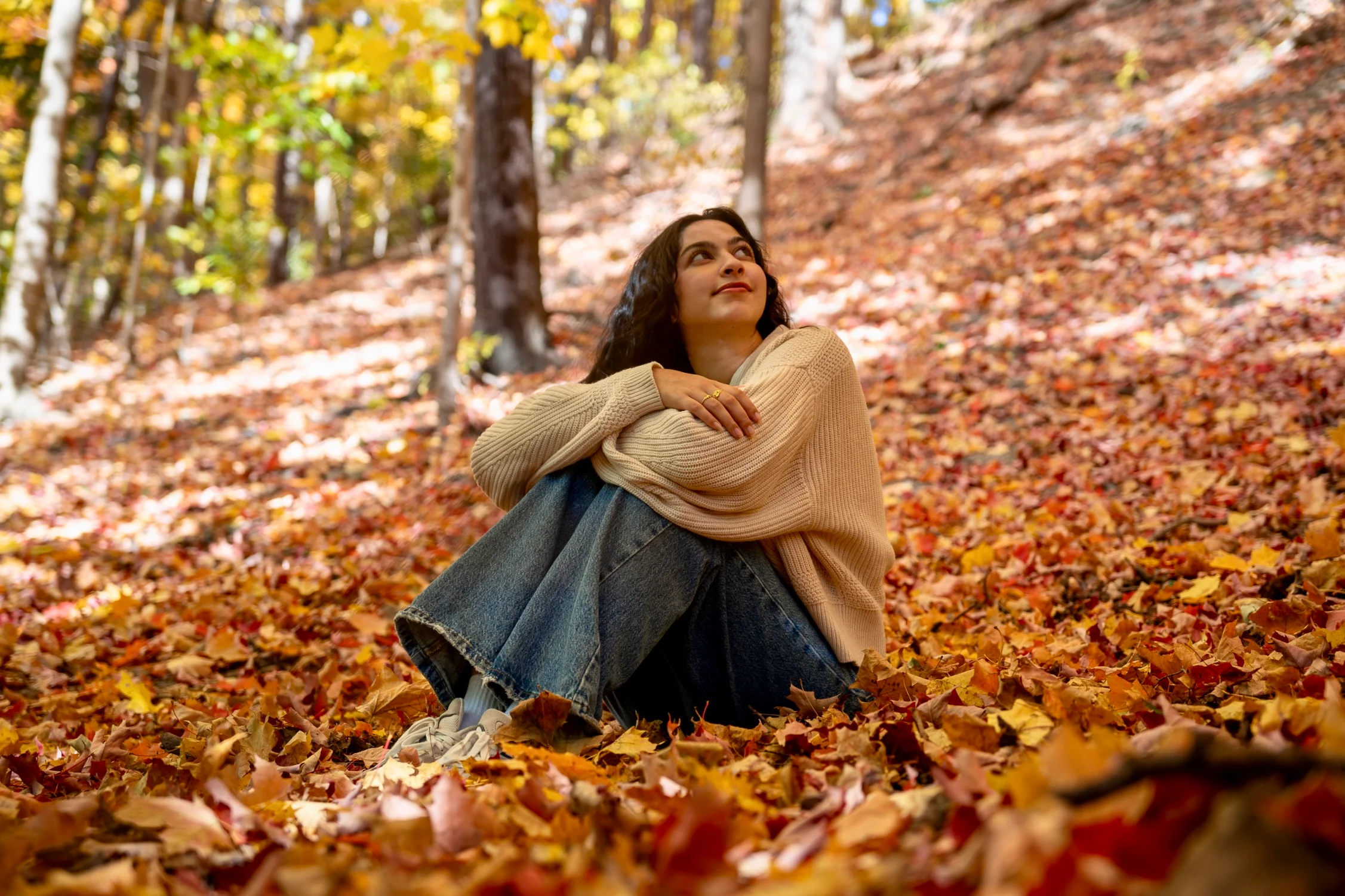 A woman sitting in a bed of autumn leaves in a warm fall forest, looking upward.