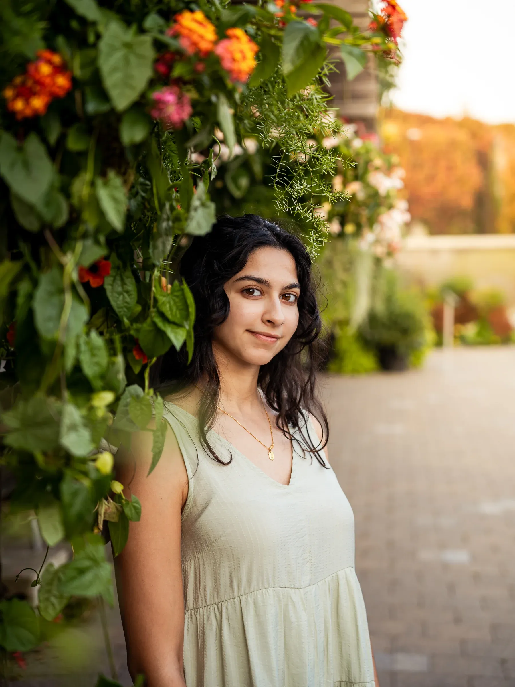 A woman in a sage dress standing beside climbing flowers at golden hour, looking calmly at the camera.