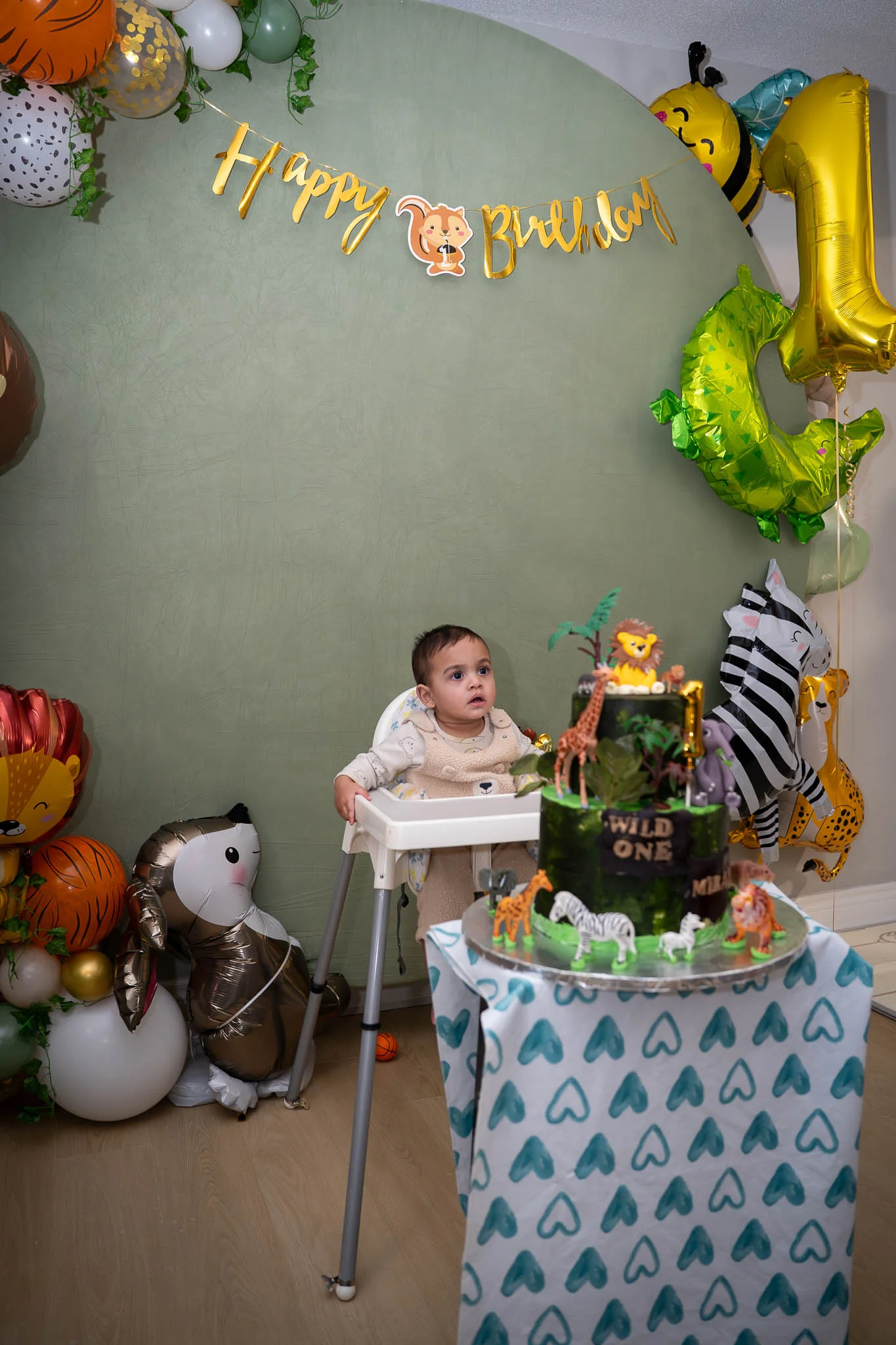A baby in a safari-themed first birthday setup with animal balloon decorations and a birthday cake.