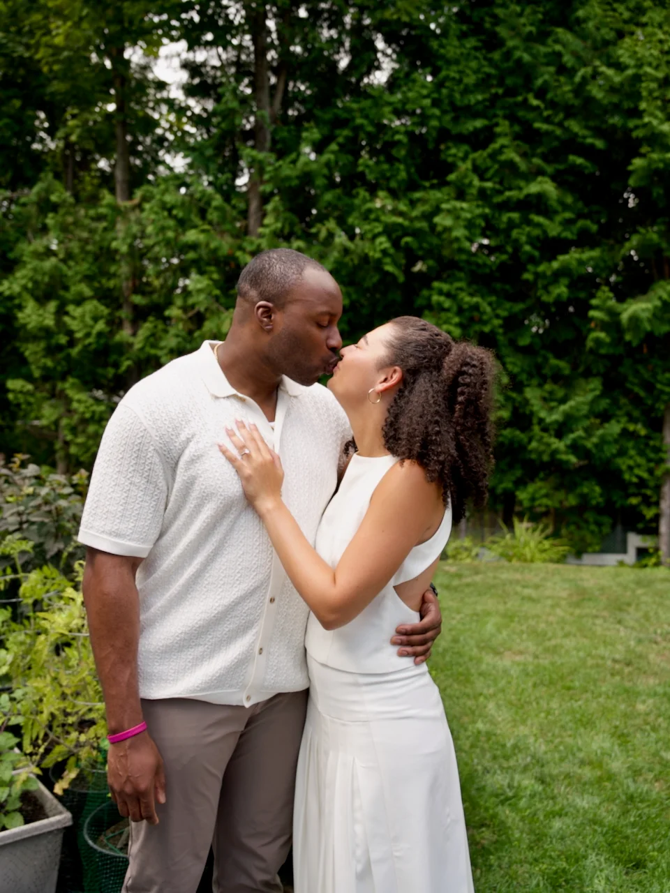 An engaged couple sharing a kiss in a green backyard, the woman in a white dress.