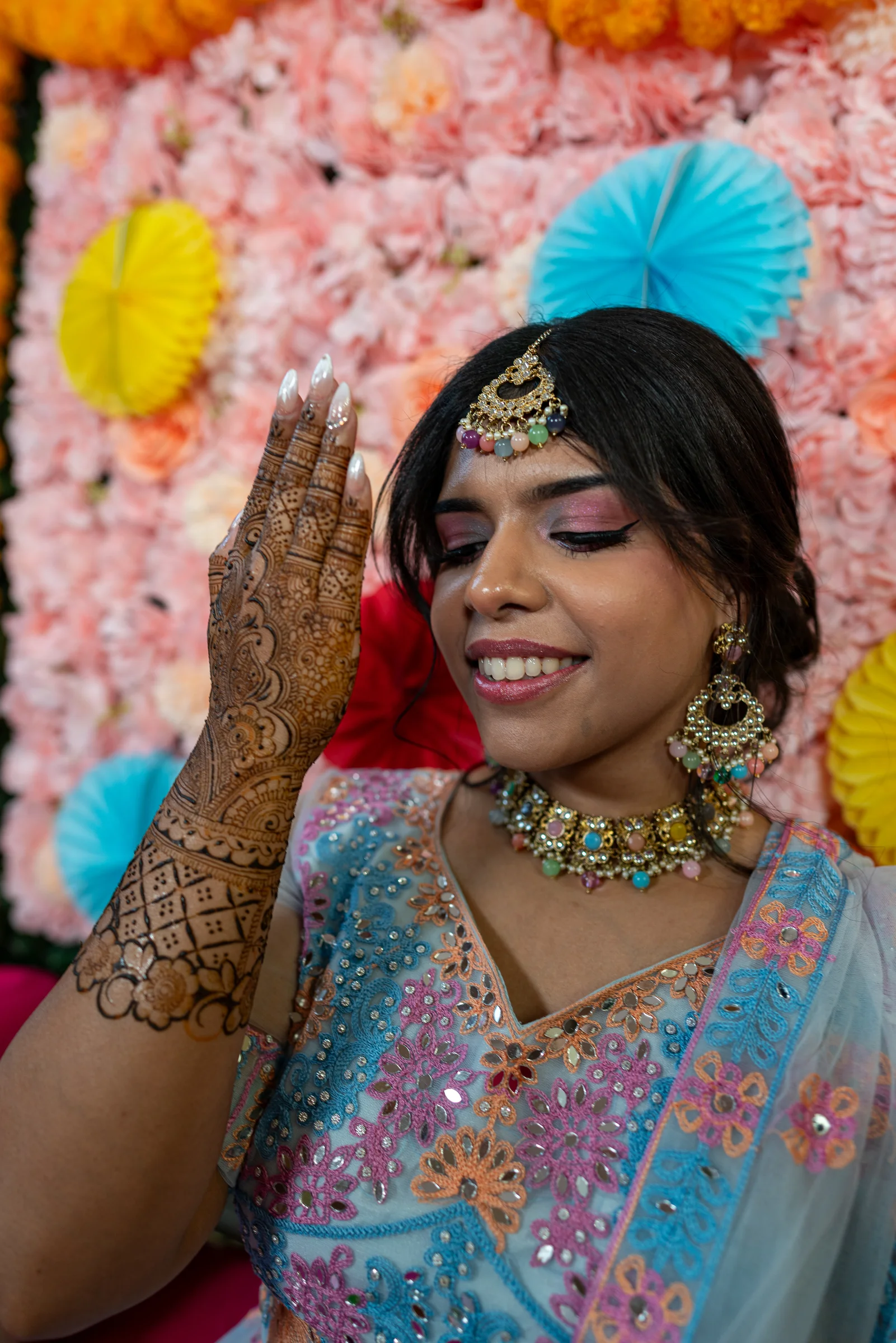 A woman with intricate henna on her hands and bridal jewellery smiling in front of a floral backdrop.