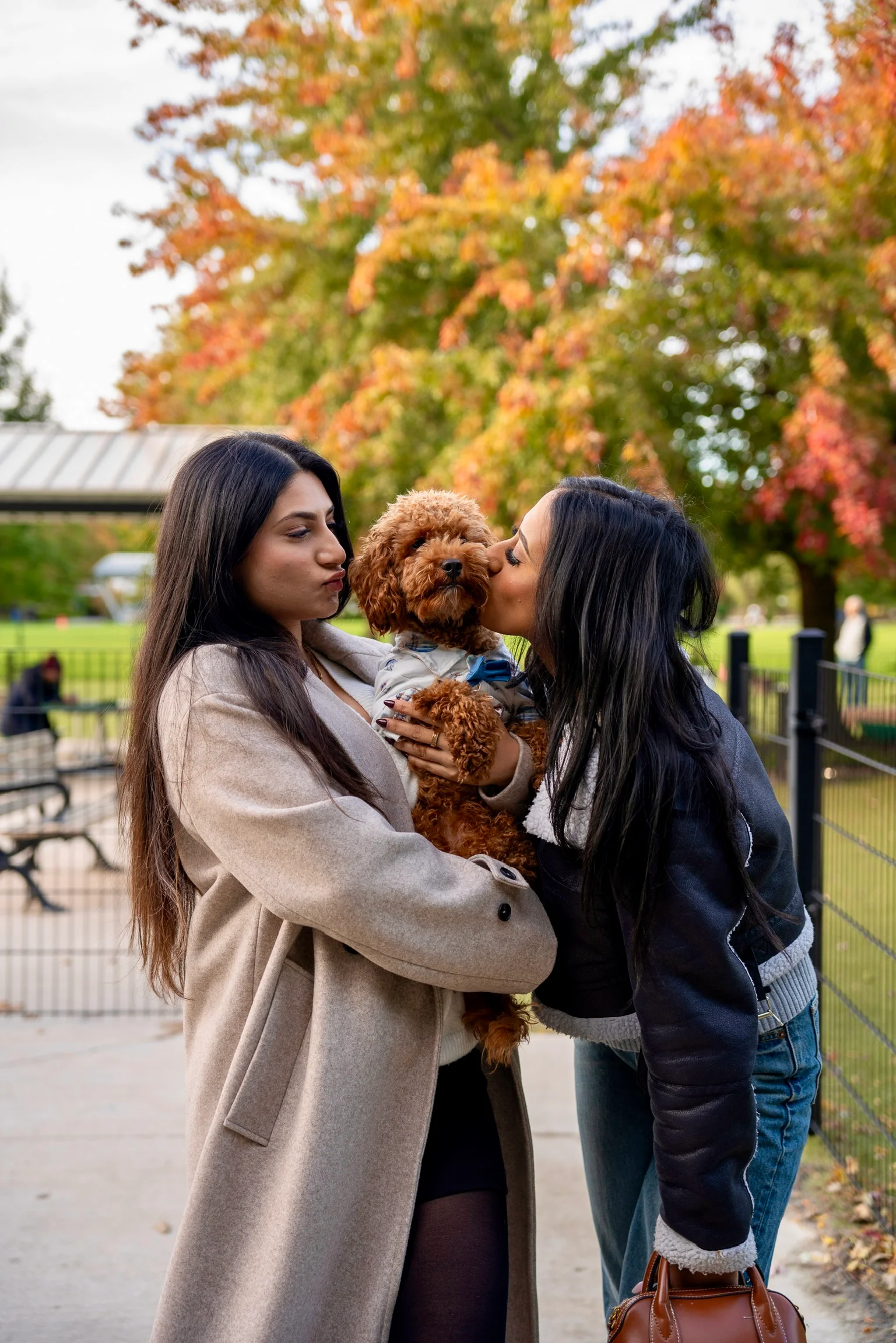 Two women outdoors among autumn trees, one holding a small apricot dog between them.